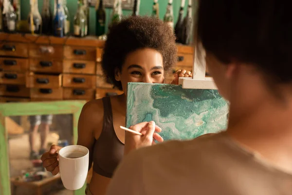 Cheerful african american woman with tea cup hiding behind easel near blurred artist — Stock Photo