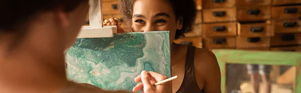 Cheerful african american woman looking from behind easel on blurred boyfriend in workshop, banner — Stock Photo