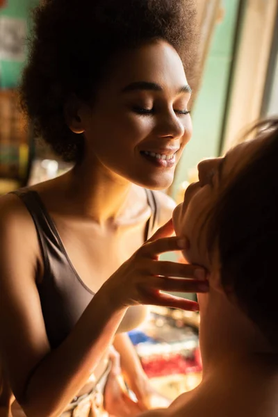 Sexy e sorridente afro-americano mulher tocando rosto de jovem artista em oficina — Fotografia de Stock