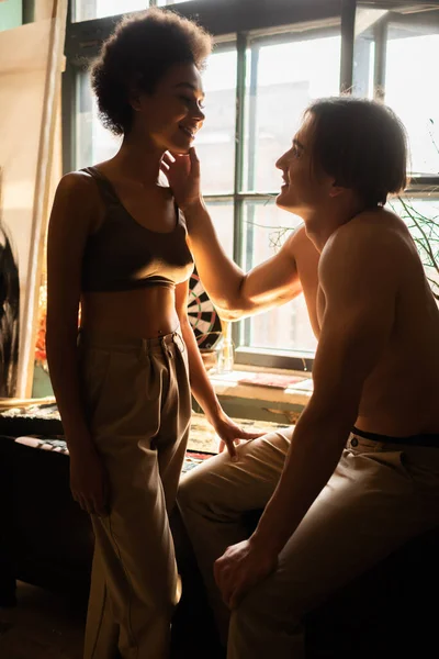 Smiling shirtless man touching face of sexy african american woman in top near window in art studio — Stock Photo