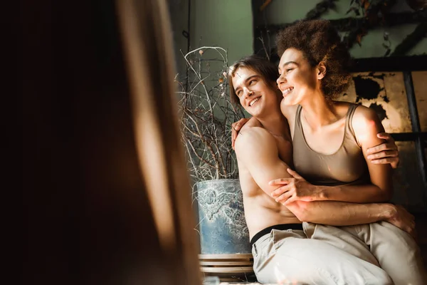 Uomo senza maglietta e allegra donna afroamericana in top abbracci e guardando lontano in studio d'arte — Foto stock