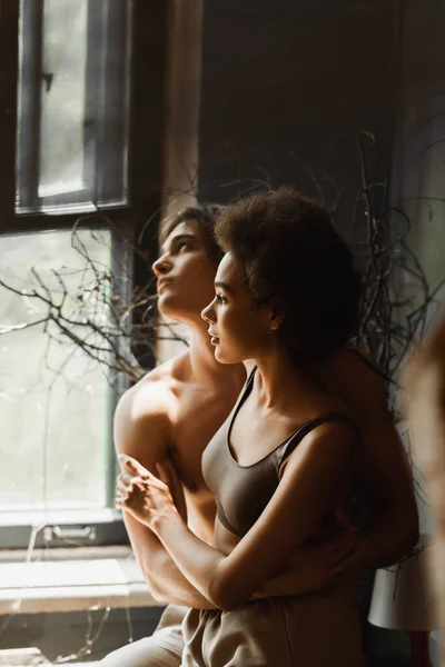 Mujer afroamericana soñadora y hombre sin camisa abrazando y mirando a través de la ventana en el taller - foto de stock