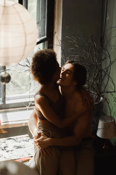 Щасливий без сорочки художник обіймає молоду афроамериканську дівчину біля вікна в художній студії — стокове фото