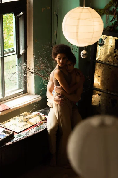 Hombre sin camisa abrazando sexy africano americano mujer mirando lejos cerca ventana en taller — Stock Photo