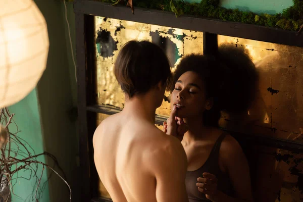 Hombre musculoso sin camisa tocando la cara de la mujer afroamericana apasionada cerca de vidrio vintage rayado - foto de stock