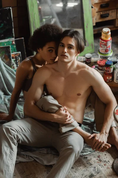 Young and passionate interracial artists sitting on floor near paints and looking at camera — Stock Photo