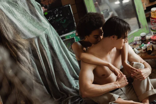 Tender african american woman kissing neck of shirtless boyfriend holding her hands on floor in art studio — Stock Photo