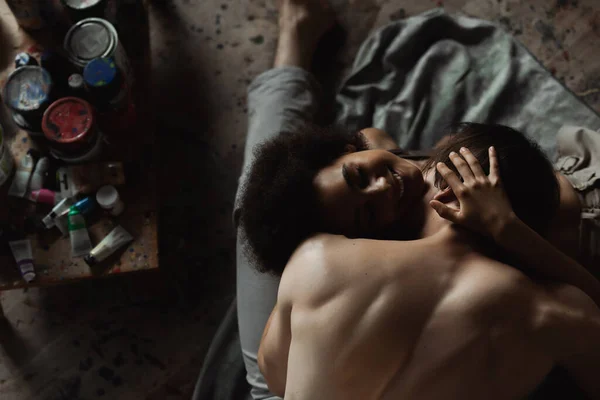 Vue du haut de l'homme musclé merdique embrasser femme afro-américaine gaie sur le sol dans un studio d'art — Photo de stock