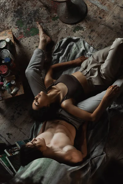 Top view of passionate african american woman in top and pants lying with closed eyes  near sexy man on floor in workshop — Stock Photo