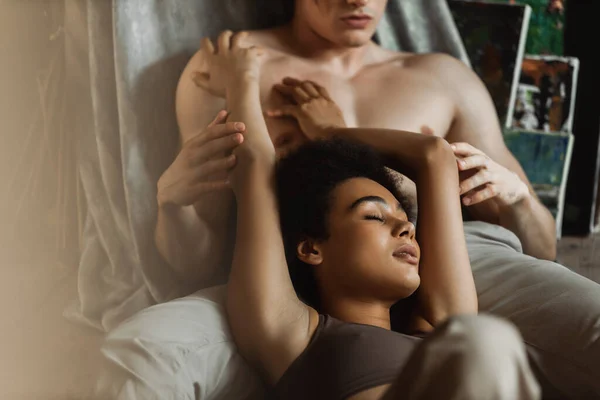 Passionate african american woman with closed eyes touching muscular shirtless man in workshop — Stock Photo