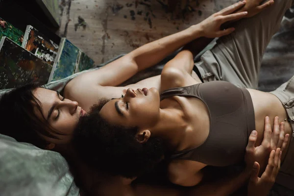 Top view of sexy african american woman lying with closed eyes near shirtless man embracing her on floor in art studio — Stock Photo