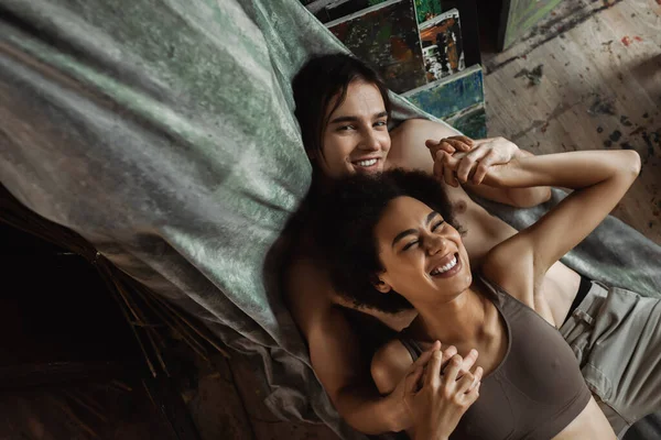 Top view of shirtless man looking at camera and holding hands with cheerful african american girlfriend — Stock Photo