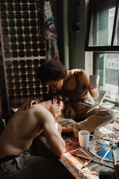 Joven hombre sin camisa apoyado en la novia afroamericana sentado en el alféizar de la ventana con una taza de té - foto de stock