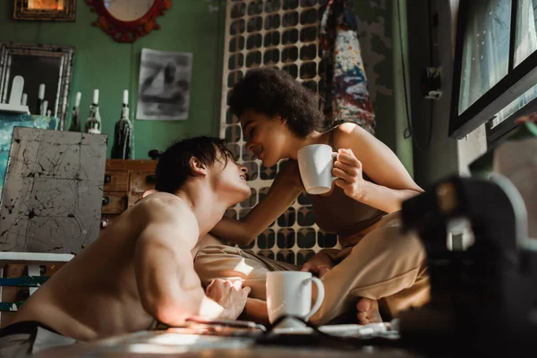 Smiling african american woman sitting on windowsill with tea cup and looking at sexy shirtless boyfriend — Stock Photo