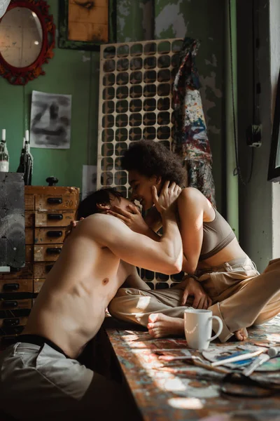 Happy african american woman sitting on windowsill and kissing with shirtless boyfriend in workshop — Stock Photo