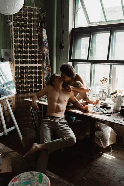 Sexy shirtless man in pants smiling near african american girlfriend sitting on windowsill — Stock Photo