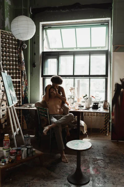 Young african american woman sitting on windowsill and embracing sexy shirtless boyfriend in pants — Stock Photo