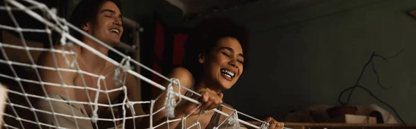 Low angle view of interracial couple laughing in mesh hammock in art studio, banner — Stock Photo