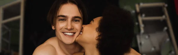 Feliz homem sem camisa olhando para a câmera, enquanto mulher afro-americana beijando ele, banner — Fotografia de Stock
