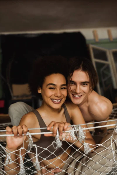 Cheerful multiethnic couple looking at camera from hammock in workshop — Stock Photo