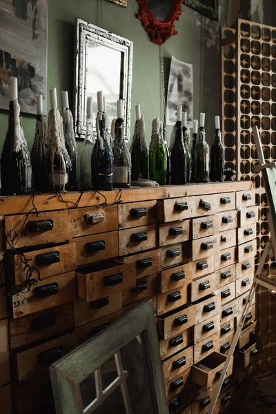 Bottles with candles on vintage rack with drawers near mirrors and paintings on wall in art studio — Stock Photo