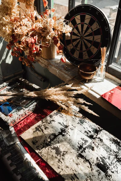 Still life of dried plants near darts target and aroma sticks on rough windowsill in sunlight — Stock Photo