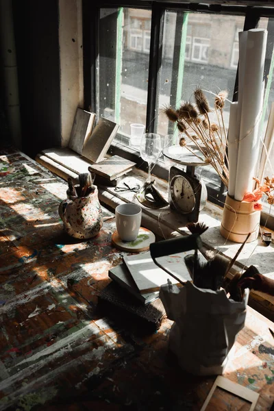 Jarrón de estuco y jarra con herramientas de arte cerca de libros y escalas vintage en alféizar de ventana áspera con manchas de pintura - foto de stock