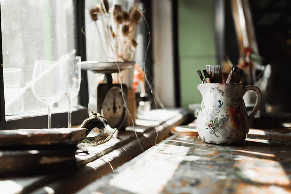 Handmade jug with paintbrushes near vintage scales and glasses on windowsill in sunlight — Stock Photo