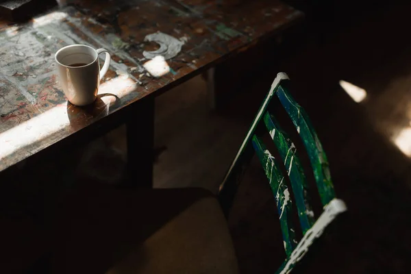 Старый деревянный стул рядом с грубым столом с пятнами краски и чашкой белого чая в художественной студии — стоковое фото