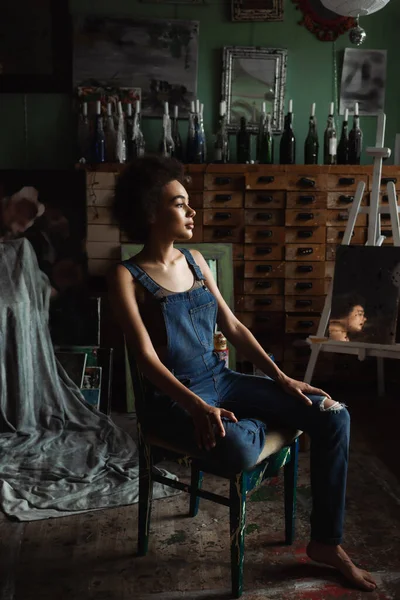 Brunette african american woman in overalls sitting in workshop with vintage decor and looking away — Stock Photo