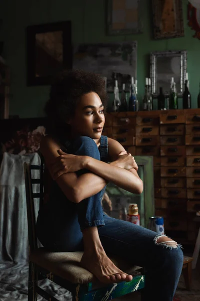 Pretty and thoughtful african american woman in overalls sitting with crossed arms in art studio — Stock Photo