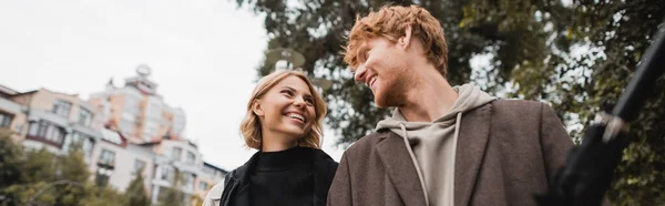 Счастливый рыжеволосый мужчина и веселая блондинка смотрят друг на друга во время прогулки в парке, баннер — стоковое фото