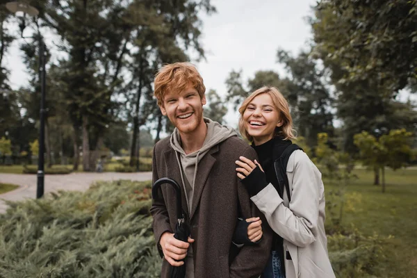 Happy blonde woman hugging cheerful redhead boyfriend with umbrella — Stock Photo