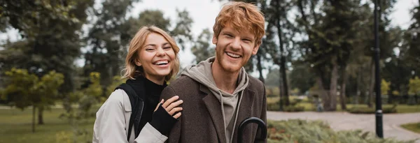 Glückliche blonde Frau umarmt fröhlichen rothaarigen Freund lächelnd im herbstlichen Park, Banner — Stockfoto