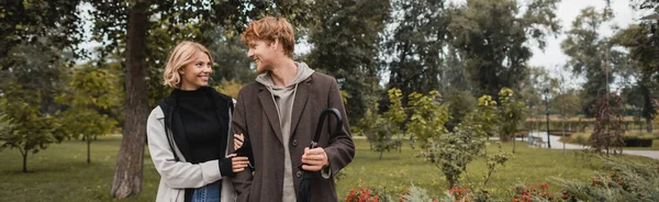 Glückliche blonde Frau umarmt fröhlichen rothaarigen Freund mit Regenschirm im Park, Banner — Stockfoto