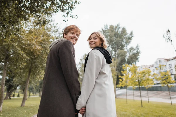 Low angle view of joyful blonde woman and cheerful redhead man holding hands while strolling in park — Stock Photo