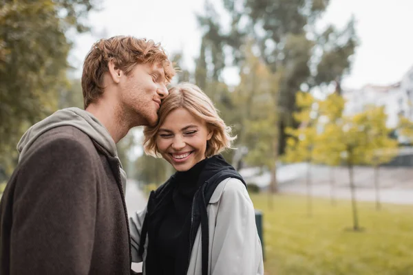 Feliz pelirroja hombre besos cabeza de rubia novia sonriendo en parque - foto de stock