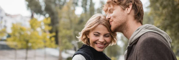 Feliz pelirroja hombre besar cabeza de rubia novia sonriendo en parque, pancarta - foto de stock