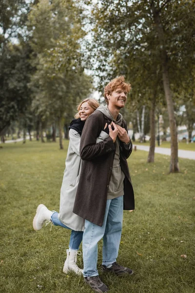 Longitud completa de la mujer joven positiva en abrigo abrazando alegre pelirroja novio en el parque otoñal - foto de stock