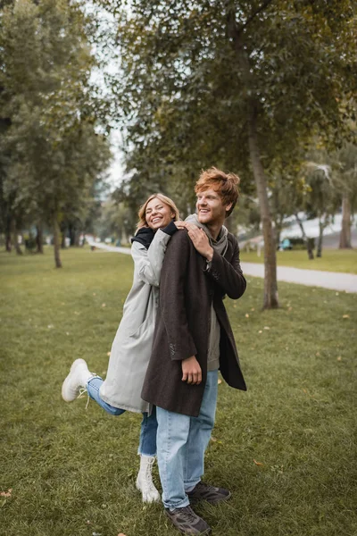 In voller Länge glückliche junge Frau im Mantel umarmt fröhlichen rothaarigen Freund im herbstlichen Park — Stockfoto