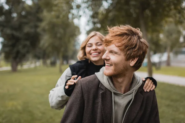 Positivo giovane donna in cappotto abbracciando allegro fidanzato rossa nel parco autunnale — Foto stock