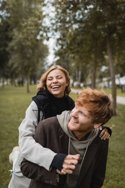 Mujer joven positiva en abrigo abrazando alegre pelirroja novio y riendo en el parque otoñal - foto de stock