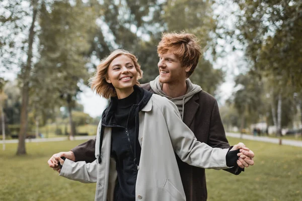 Joven positivo en abrigo cogido de la mano con alegre novia rubia en el parque otoñal - foto de stock