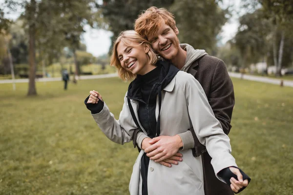 Pleased young man in coat hugging cheerful blonde girlfriend in autumnal park — Stock Photo