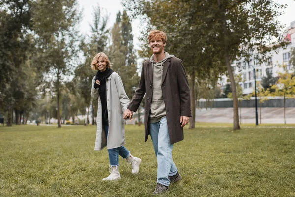 Full length of happy young man in coat holding hands with cheerful girlfriend while walking in autumnal park — Stock Photo