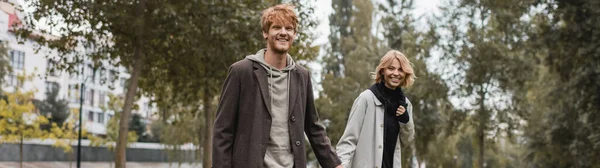 Jovem feliz no casaco de mãos dadas com namorada alegre enquanto caminha no parque outonal, banner — Fotografia de Stock