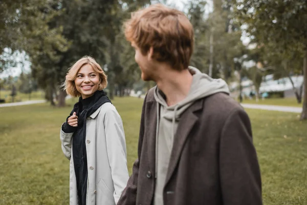 Pleased young woman in coat looking at blurred redhead boyfriend in autumnal park — Stock Photo