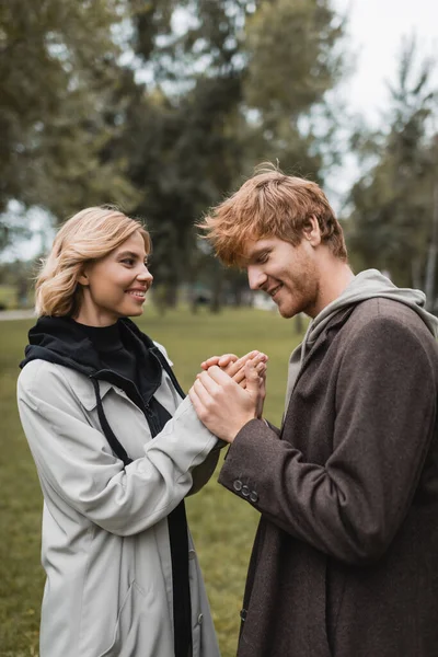 Cuidado joven en abrigo calentando manos de alegre novia en el parque otoñal - foto de stock