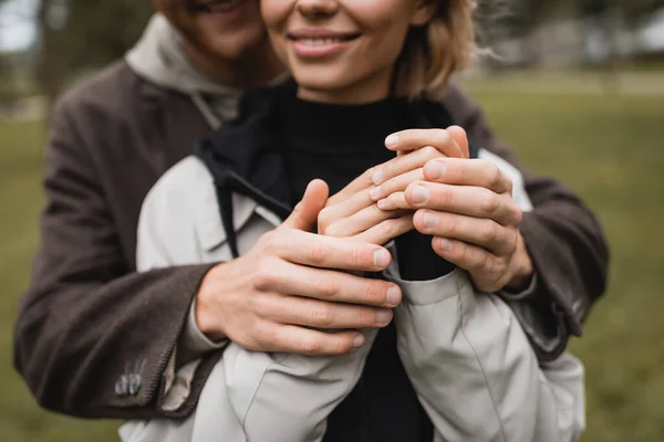 Cropped view of caring young man in coat warming up hands of cheerful girlfriend in autumnal park — Stock Photo