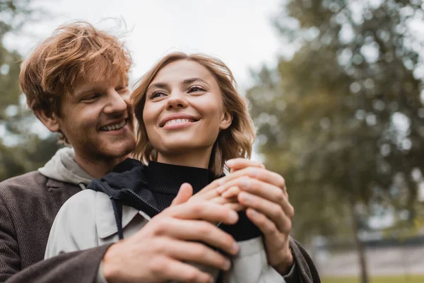Low angle view of caring young man in coat warming up hands of cheerful girlfriend in autumnal park — Stock Photo